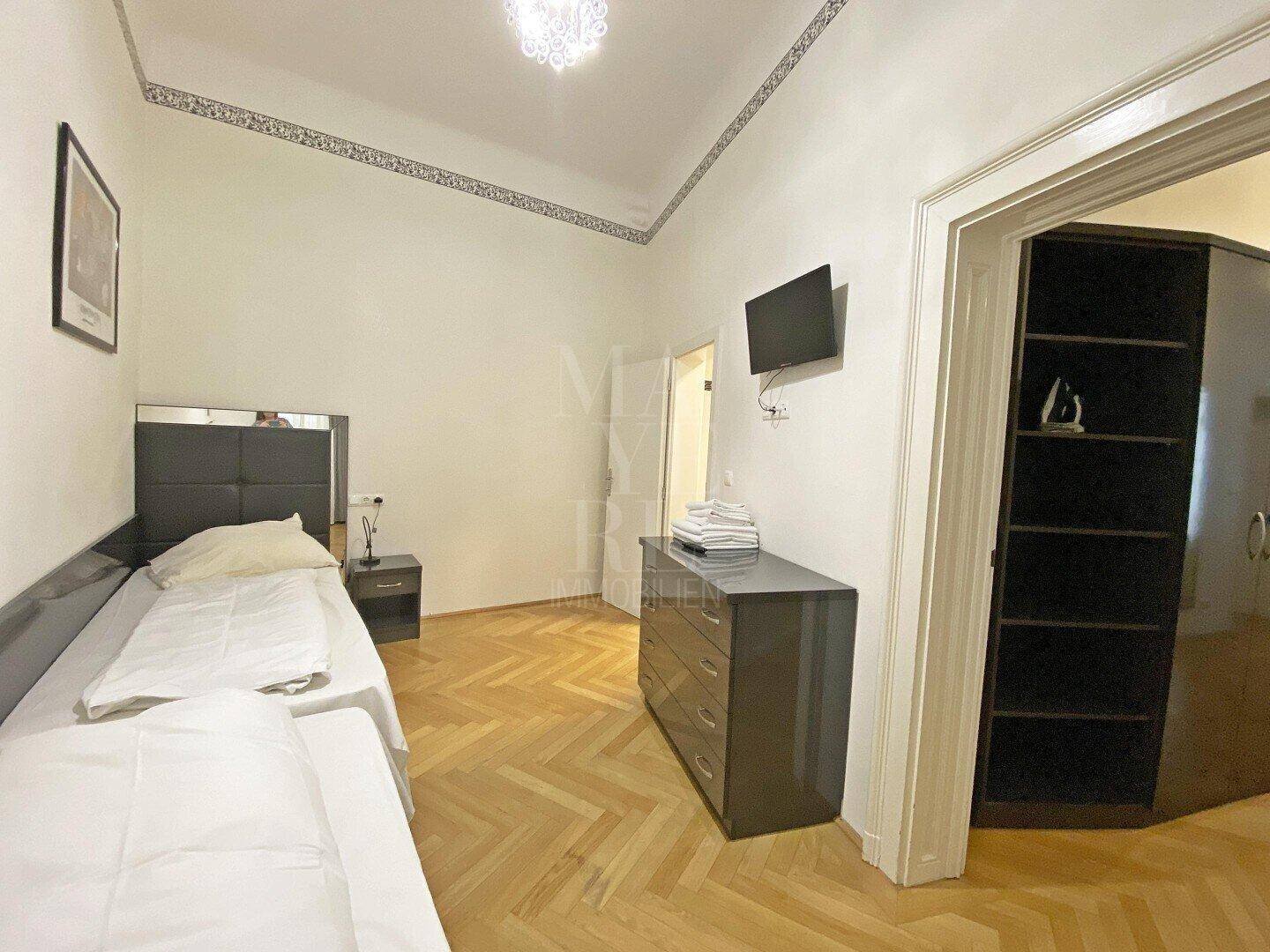 Helles Schlafzimmer mit zwei Einzelbetten, Fischgrätparkett und Fernseher.