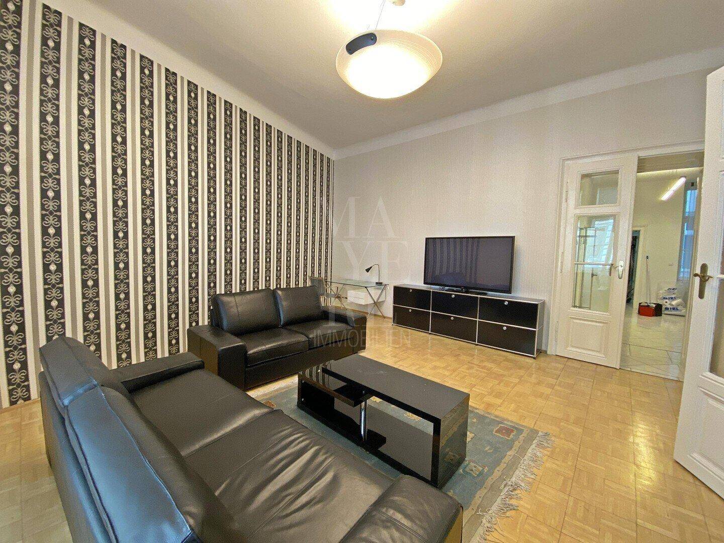 Helles Wohnzimmer mit schwarzen Ledersofas, Fernseher und auffälliger Streifentapete.