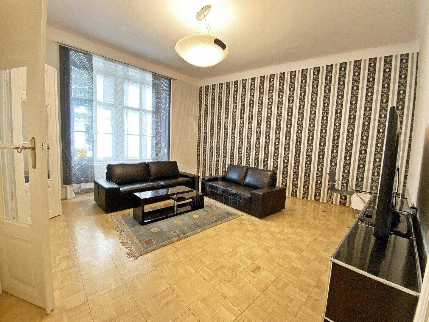 Wohnzimmer mit zwei schwarzen Sofas, Parkettboden und großen Fenstern mit Vorhängen.