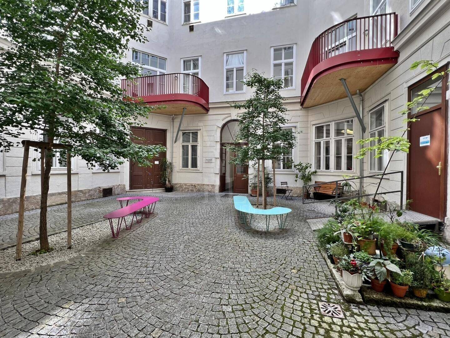 Ein charmanter Innenhof mit Kopfsteinpflaster, bunten Sitzbänken und begrünten Bereichen, umgeben von einem Altbau.