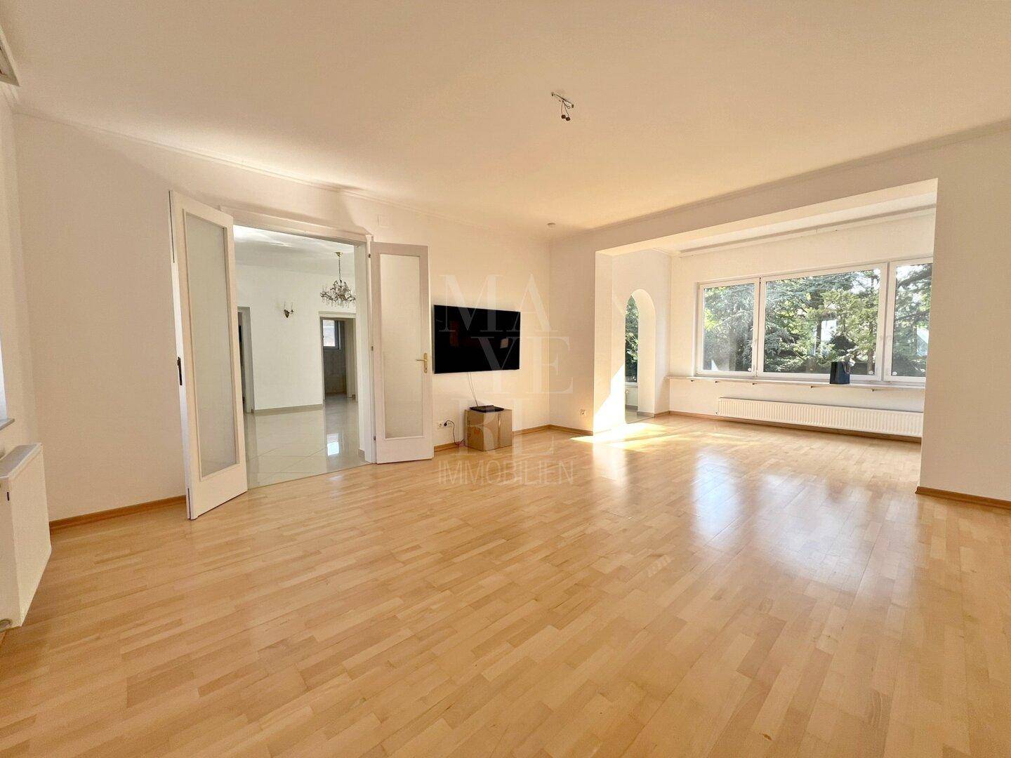 Geräumiges Wohnzimmer mit hellem Parkettboden und großen Fenstern, die viel Tageslicht hereinlassen.