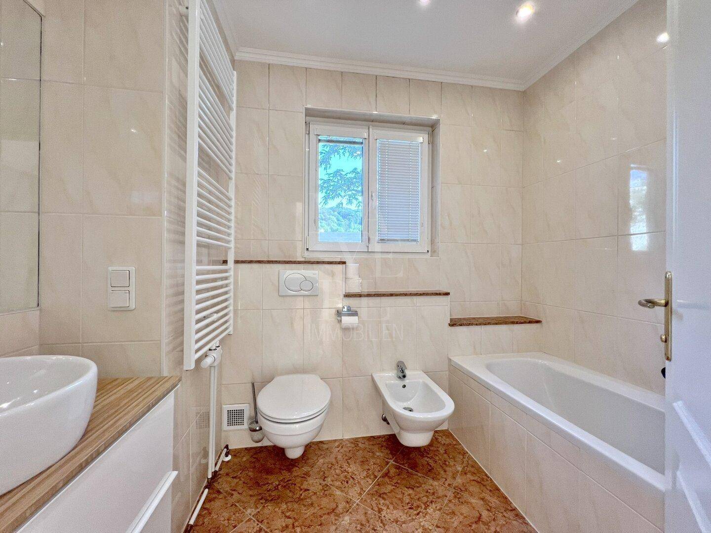 Helles Badezimmer mit Badewanne, Bidet, Toilette und Fenster, ausgestattet mit Handtuchheizkörper.