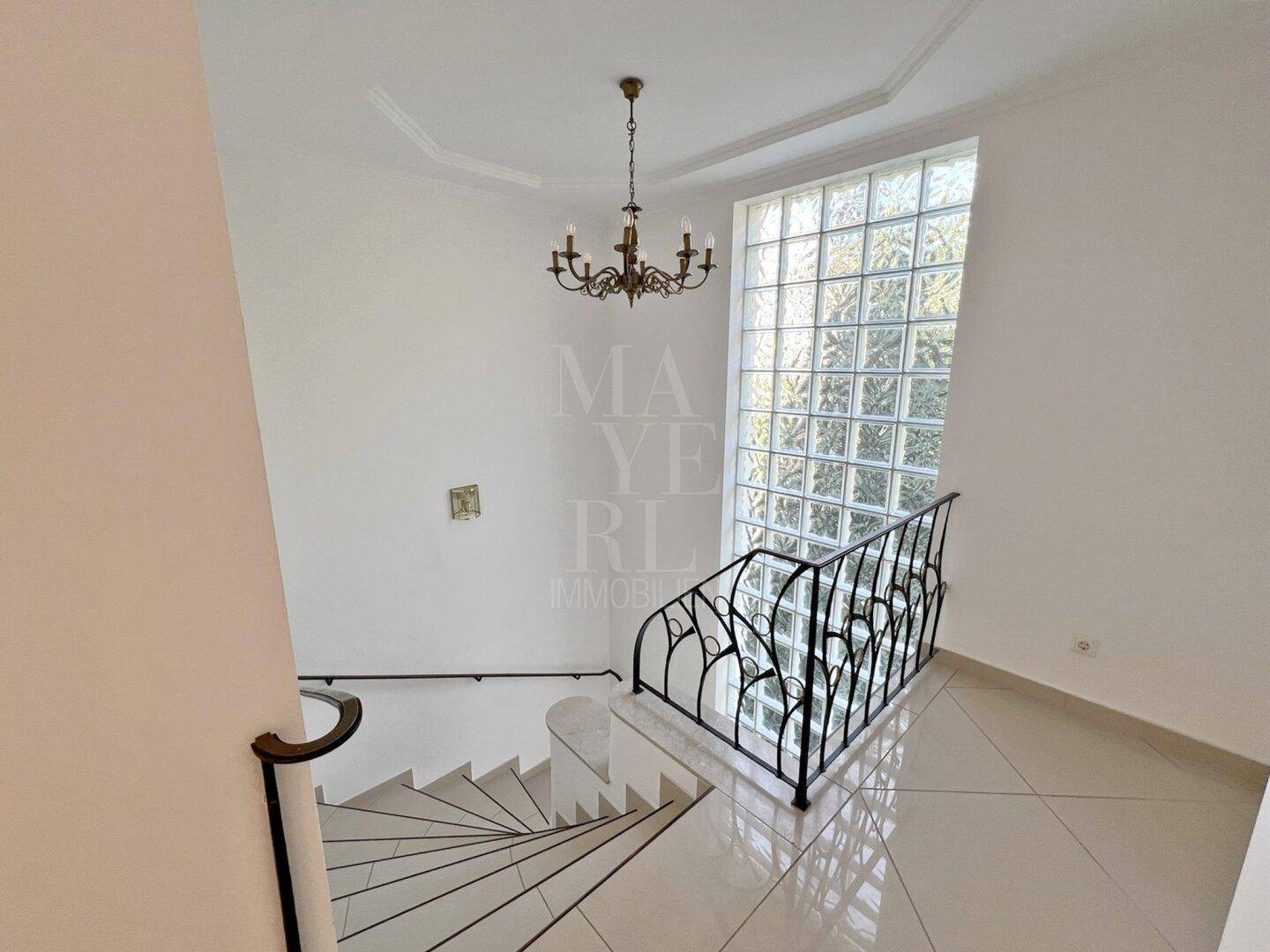 Elegantes Treppenhaus mit schmiedeeisernem Geländer und einer Glasbausteinwand, beleuchtet von einem Kronleuchter.