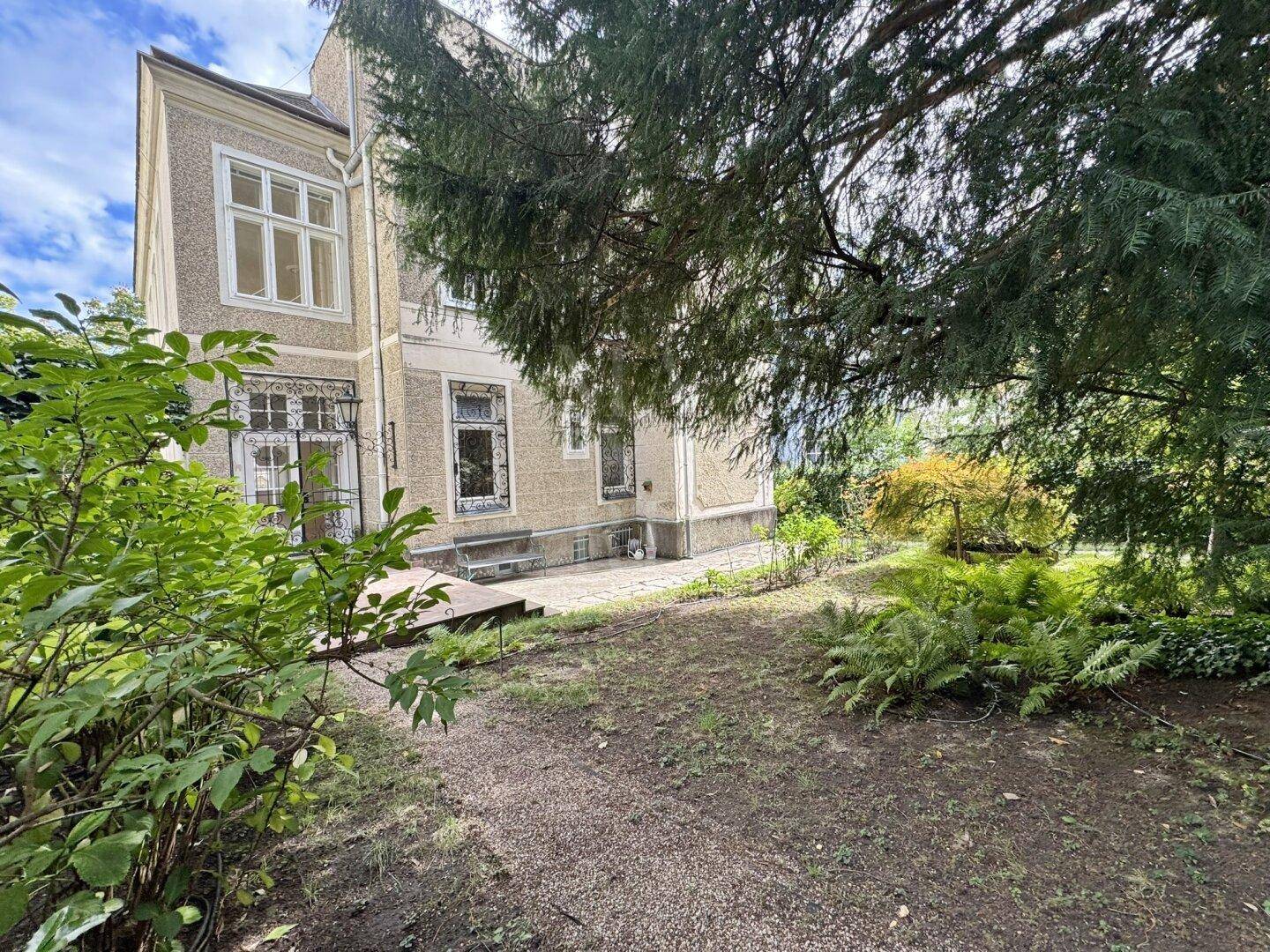 Historisches Gebäude mit gepflegtem Gartenbereich und altem Baumbestand unter bewölktem Himmel.