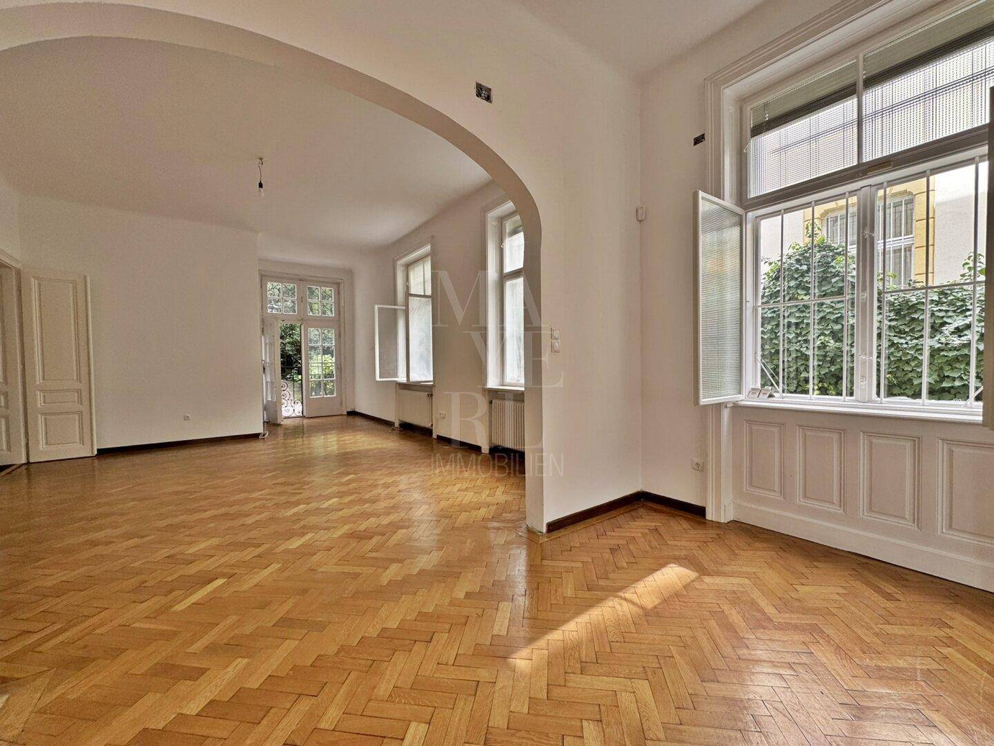 Helles Wohnzimmer mit Fischgrätparkett, hohen Decken und großen Fenstern zum Gartenbereich.