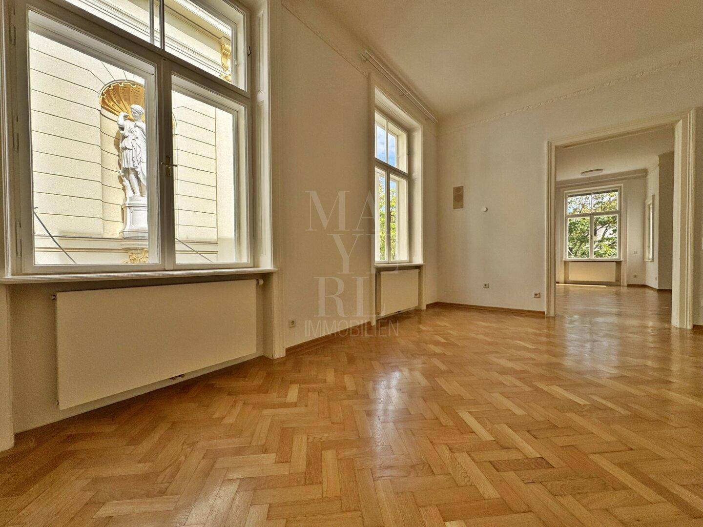 Geräumiges Wohnzimmer mit Fischgrätparkett, Stuckdecke und Blick auf eine Außenstatue durch das Fenster.
