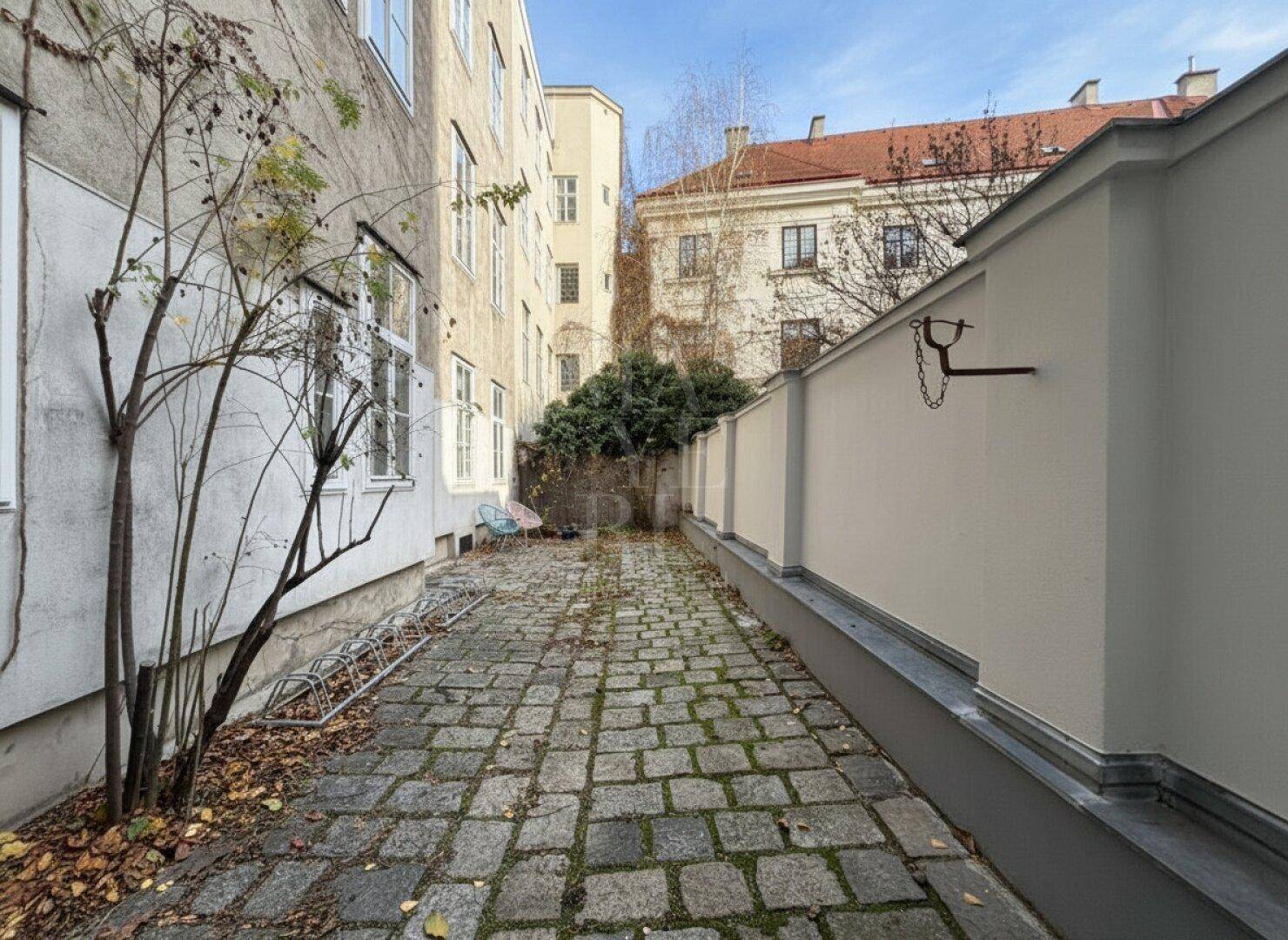 Gepflasterter Hinterhof mit Bäumen und Sträuchern, der zu einem kleinen Gartenbereich führt.