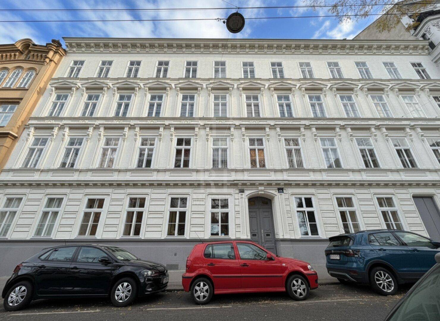 Klassische Altbaufassade mit vielen Fenstern und geparkten Autos auf der Straße davor.