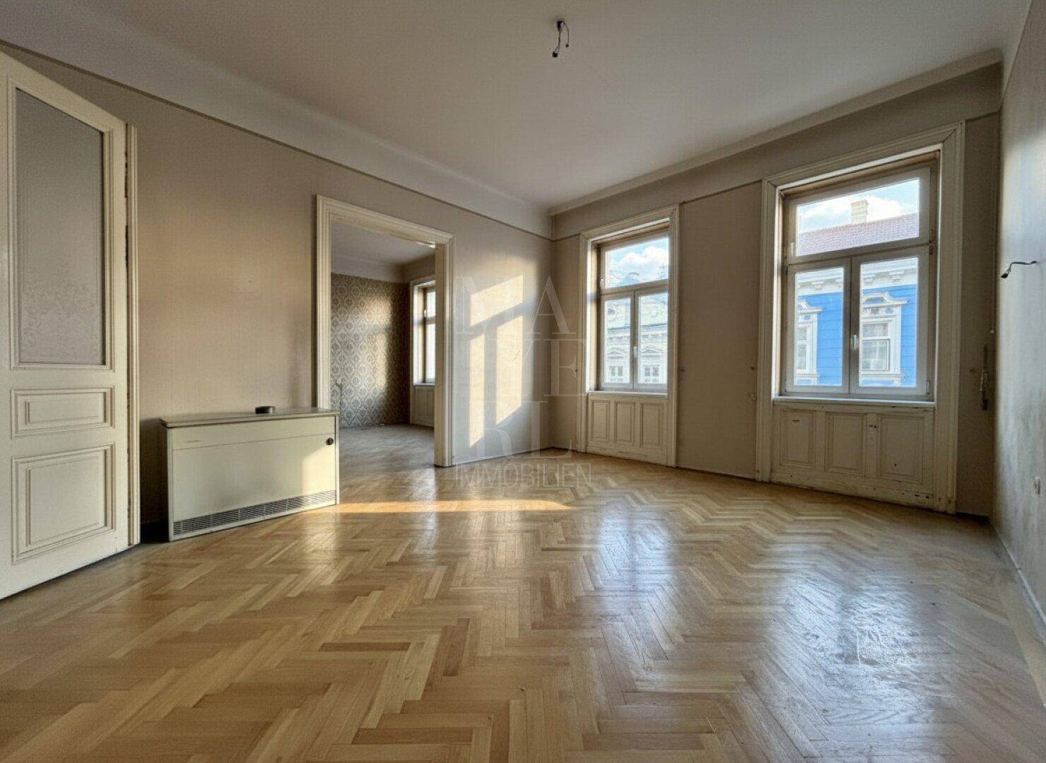 Geräumiges Zimmer mit Fischgrätparkett und hohen Fenstern, das auf eine Renovierung wartet.
