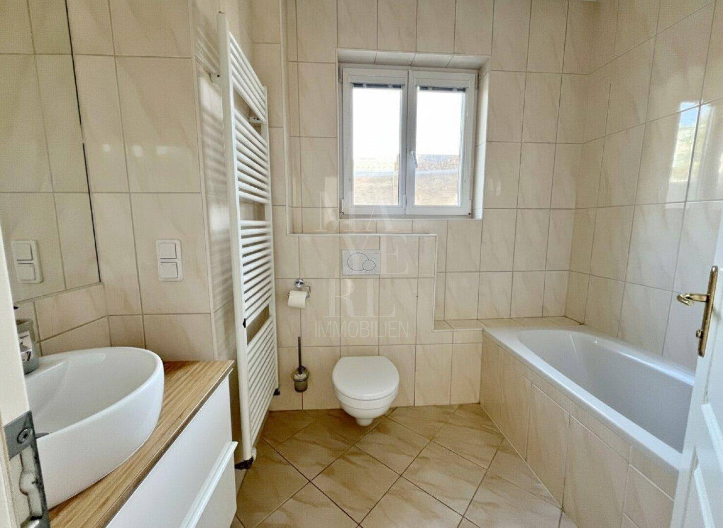 Badezimmer mit beigefarbenen Fliesen, Badewanne, WC, Einzelwaschbecken und beheiztem Handtuchhalter.