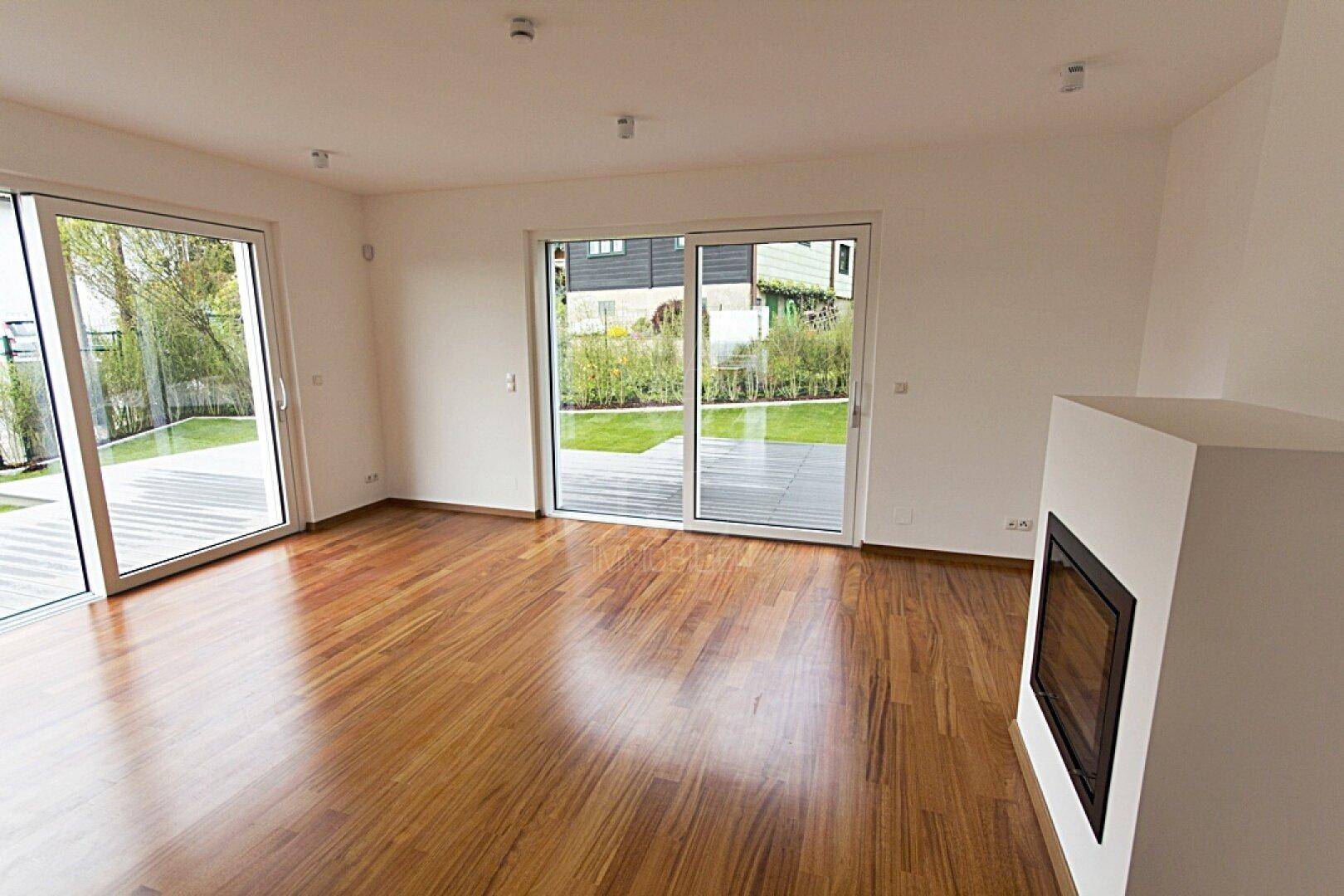 Geräumiges Wohnzimmer mit glänzendem Holzboden, Kamin und Blick auf den grünen Garten.