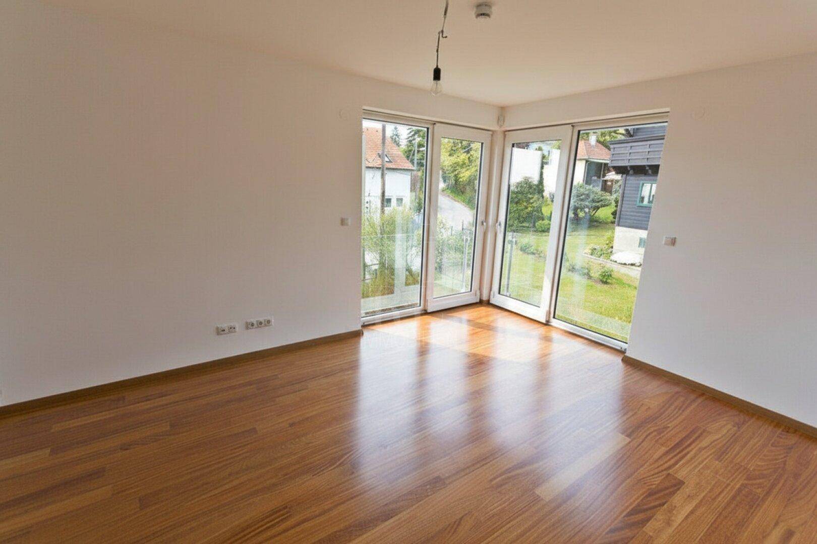 Heller Wohnraum mit großen Eckfenstern, die viel Tageslicht hereinlassen und einen Blick ins Grüne bieten.