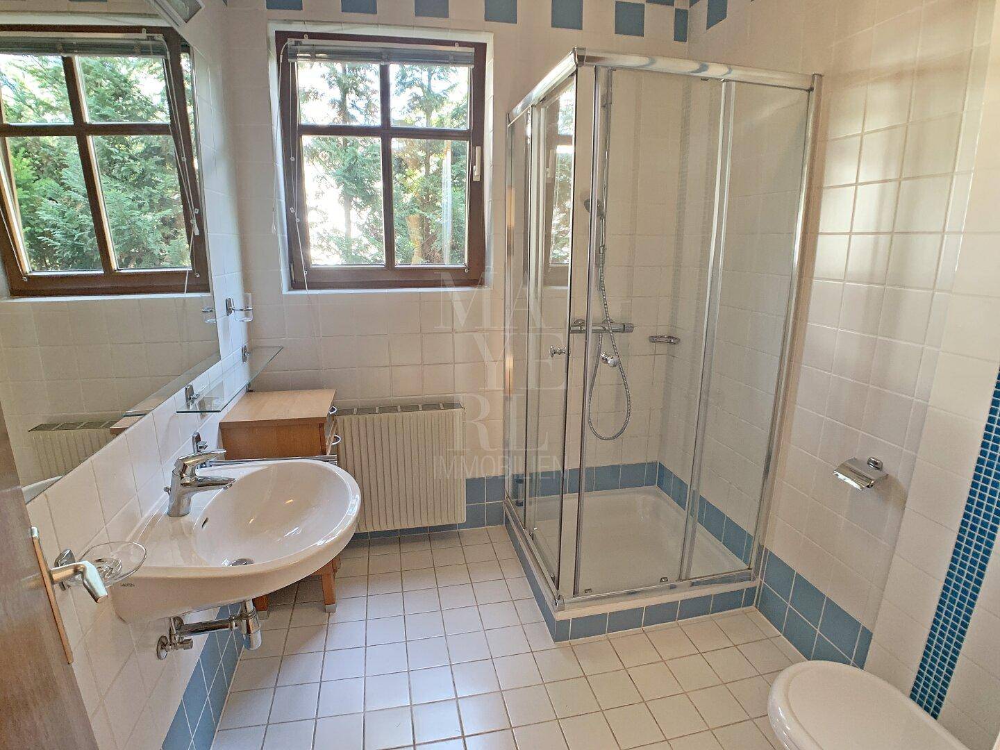 Badezimmer mit Duschkabine, Einzelwaschbecken und Fenster, hell gefliest in Weiß und Blau.