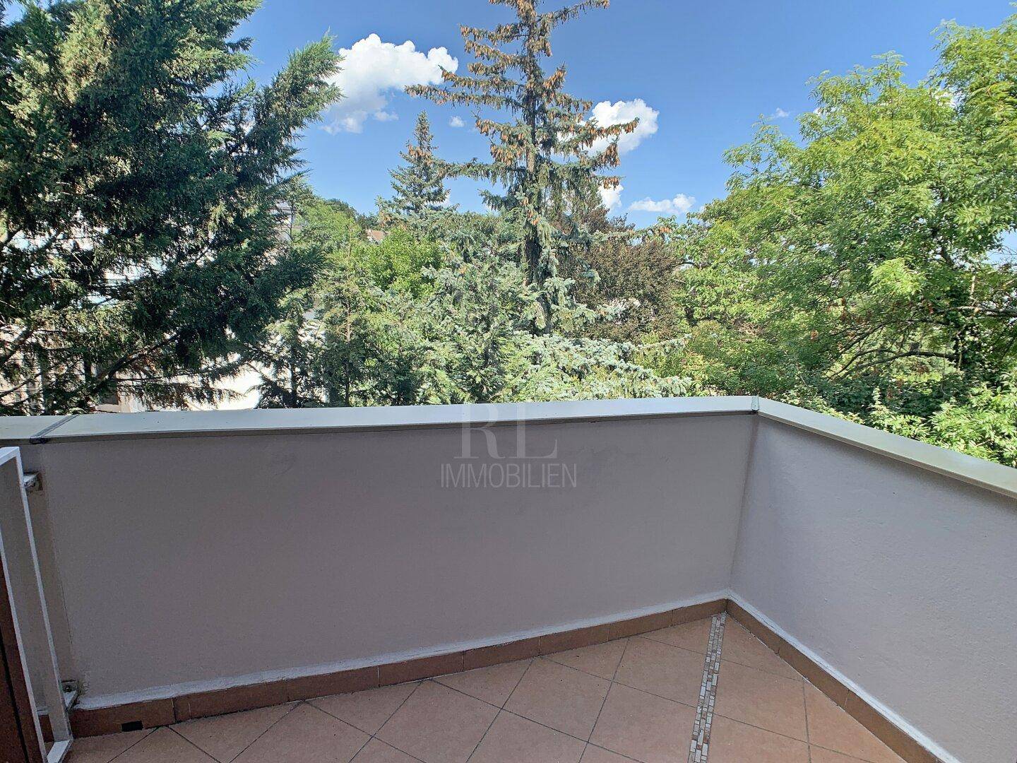 Kleiner Balkon mit Fliesenboden und Blick auf üppiges Grün und Bäume.