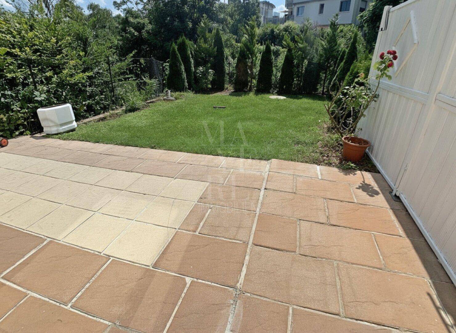 Gepflasterte Terrasse grenzt an einen kleinen, gepflegten Rasengarten mit Hecken und Bäumen.