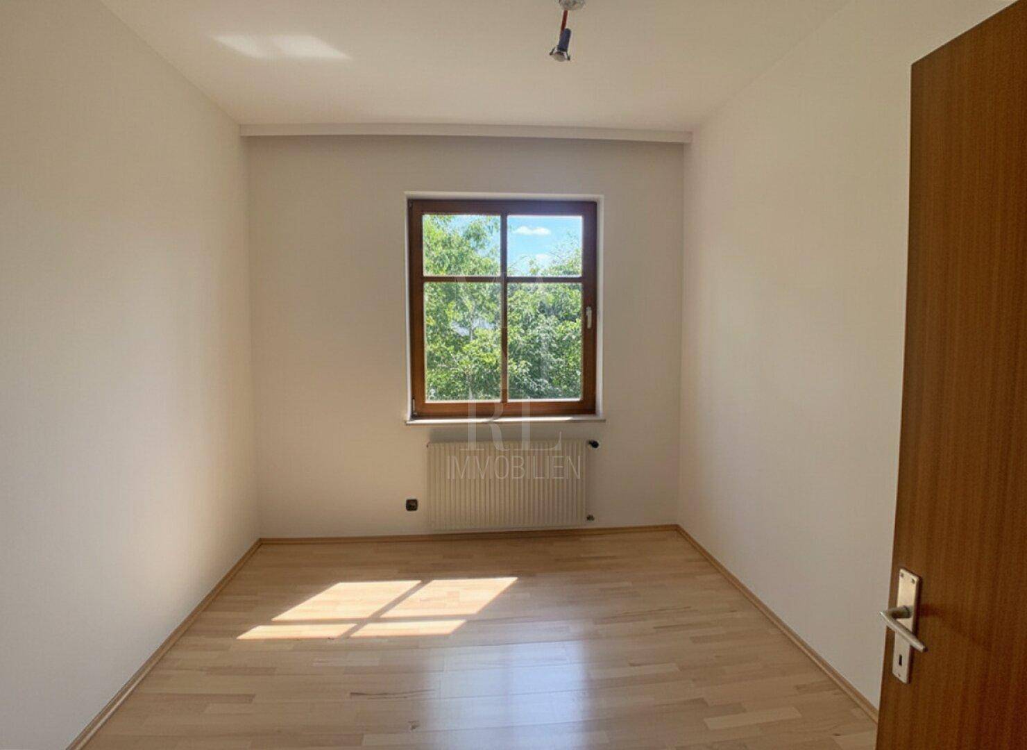 Helles Schlafzimmer mit Parkettboden und großem Fenster, das viel Tageslicht hereinlässt.