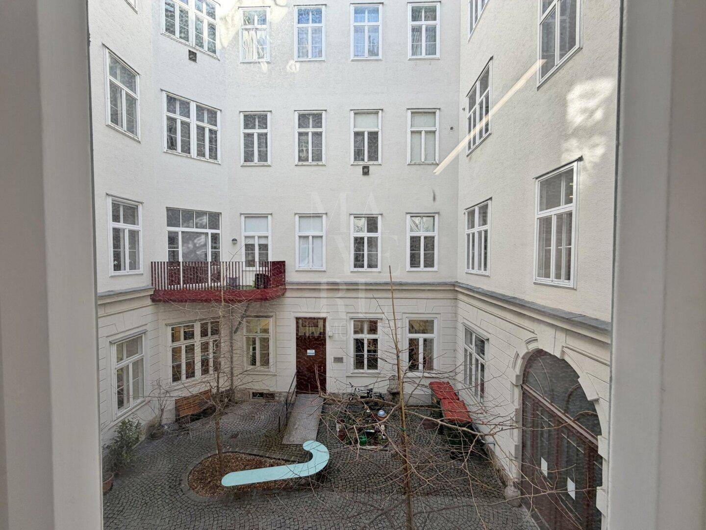 Blick aus dem Fenster auf einen gepflasterten Innenhof mit Bäumen und Balkonen.