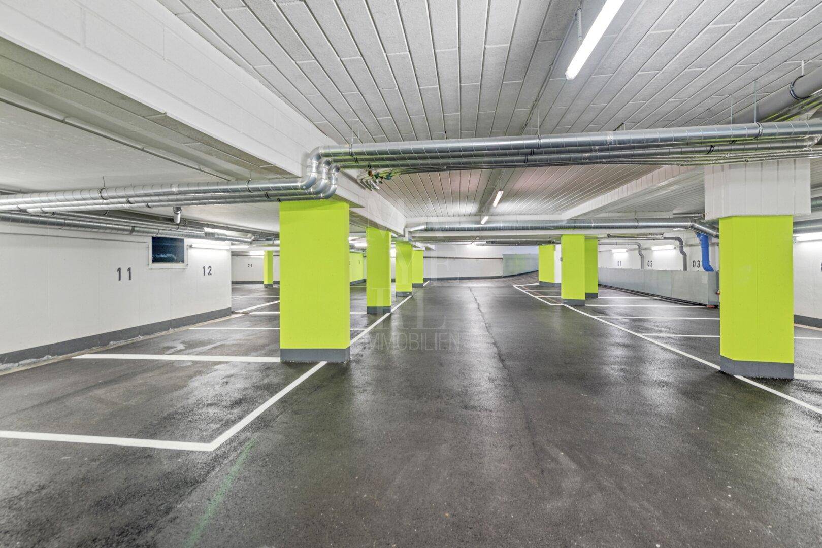 Geräumige Tiefgarage mit vielen Stellplätzen, sichtbaren Lüftungsrohren und moderner Beleuchtung.