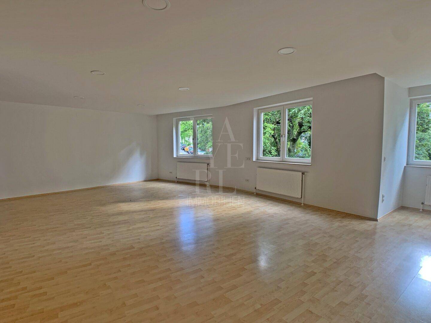 Lichtdurchflutetes Wohnzimmer mit mehreren Fenstern, hellem Parkettboden und Blick ins Grüne.