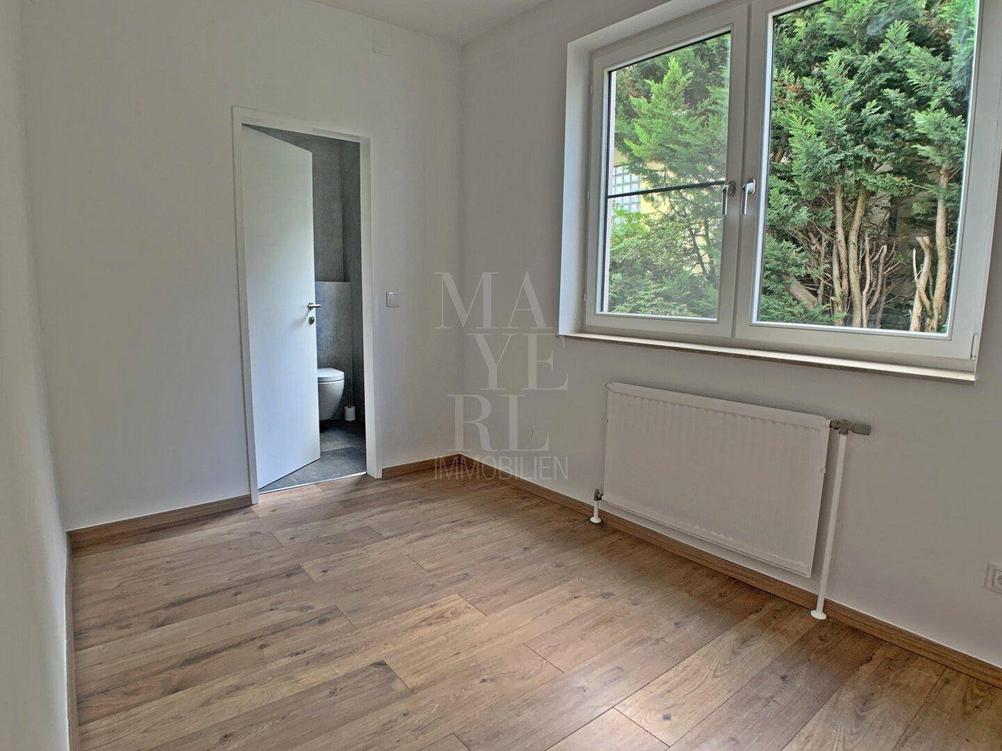Schlafzimmer mit Holzboden, Fenster und angrenzendem Badezimmerzugang.