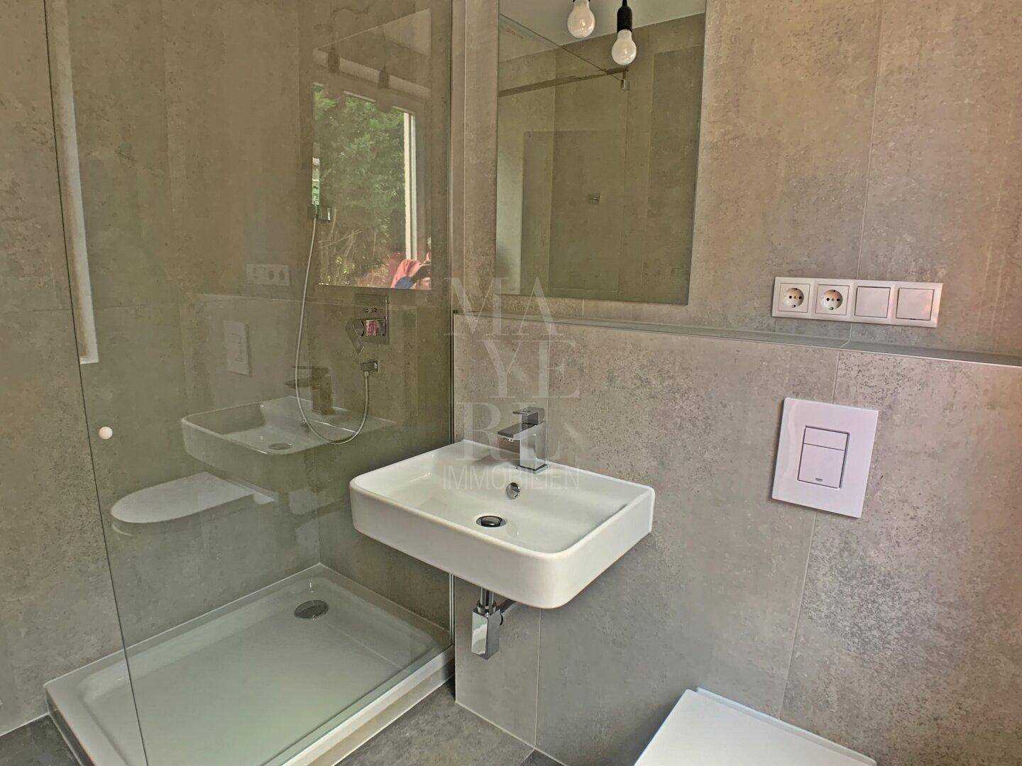 Modernes Badezimmer mit ebenerdiger Dusche, Waschbecken und grauen Fliesen an Wänden und Boden.