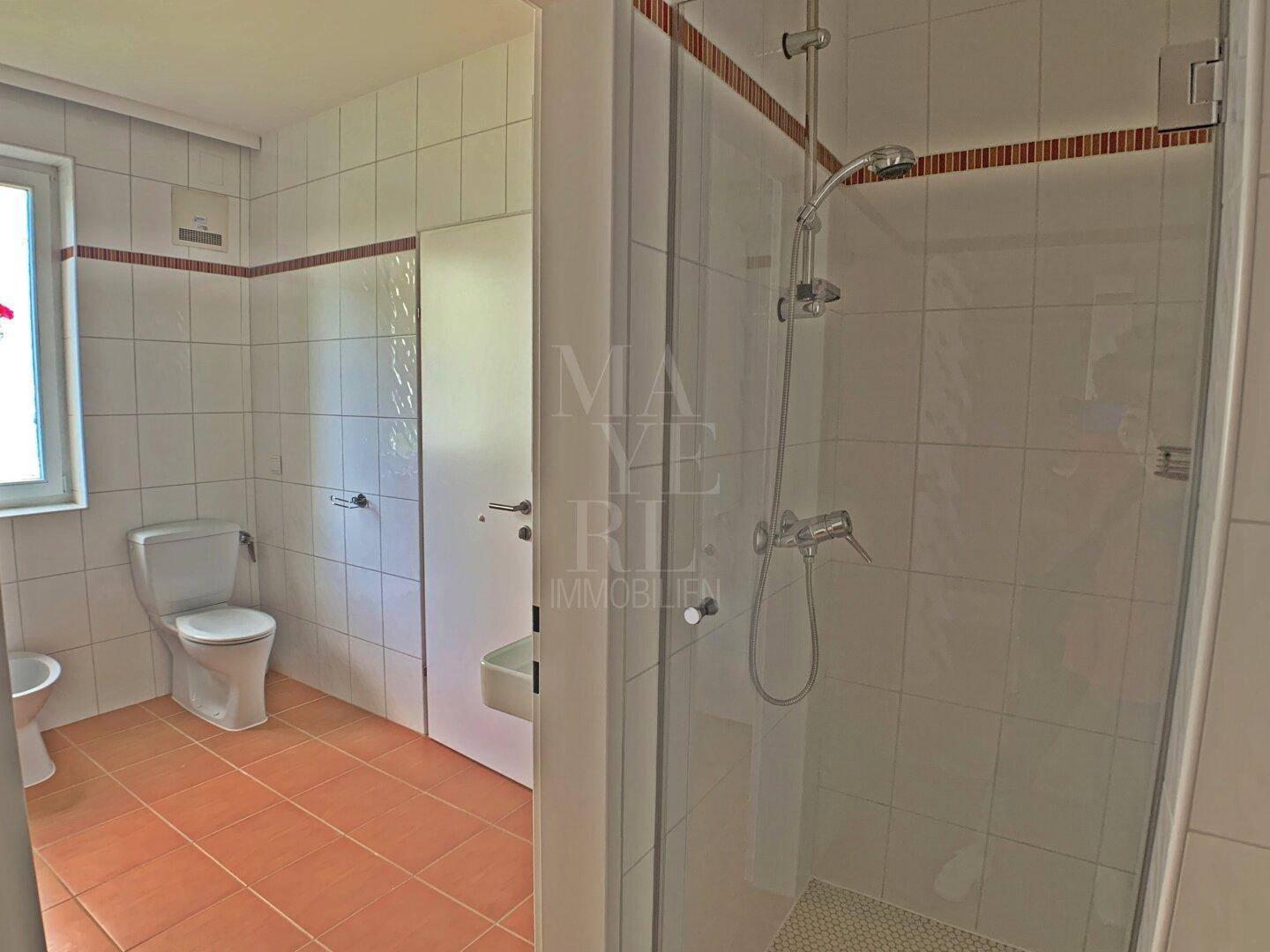 Helles Badezimmer mit Dusche, Toilette und Fenster, ausgestattet mit weißen Fliesen.