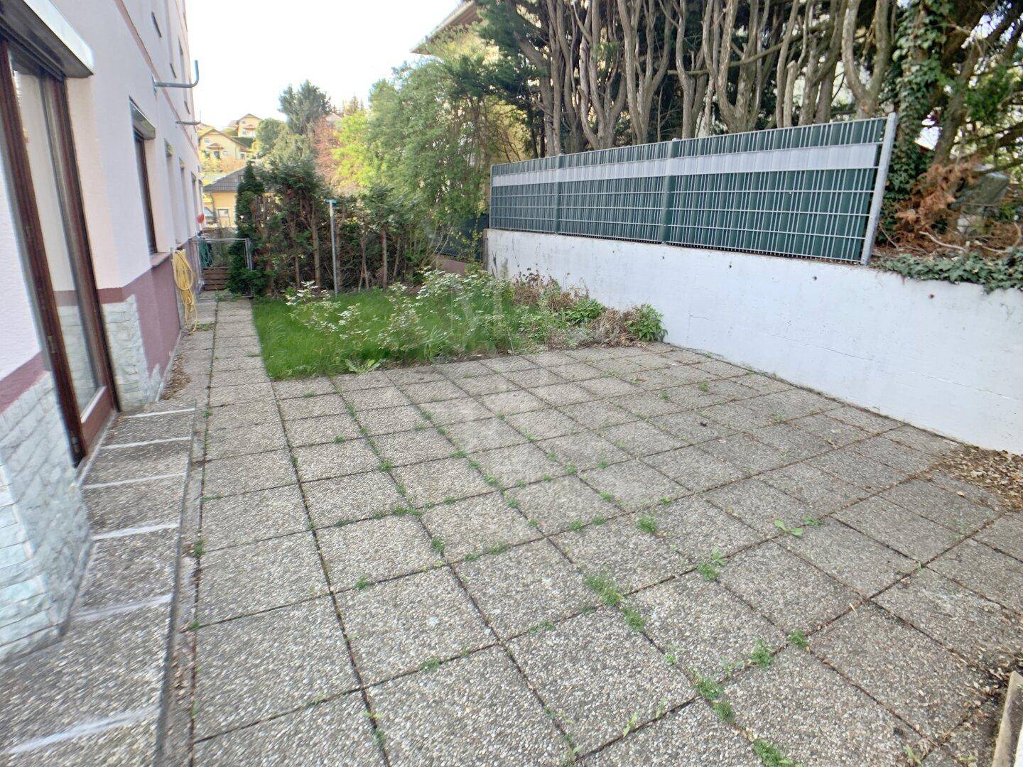 Gepflasterte Terrasse mit Rasenfläche und grüner Bepflanzung im Außenbereich.