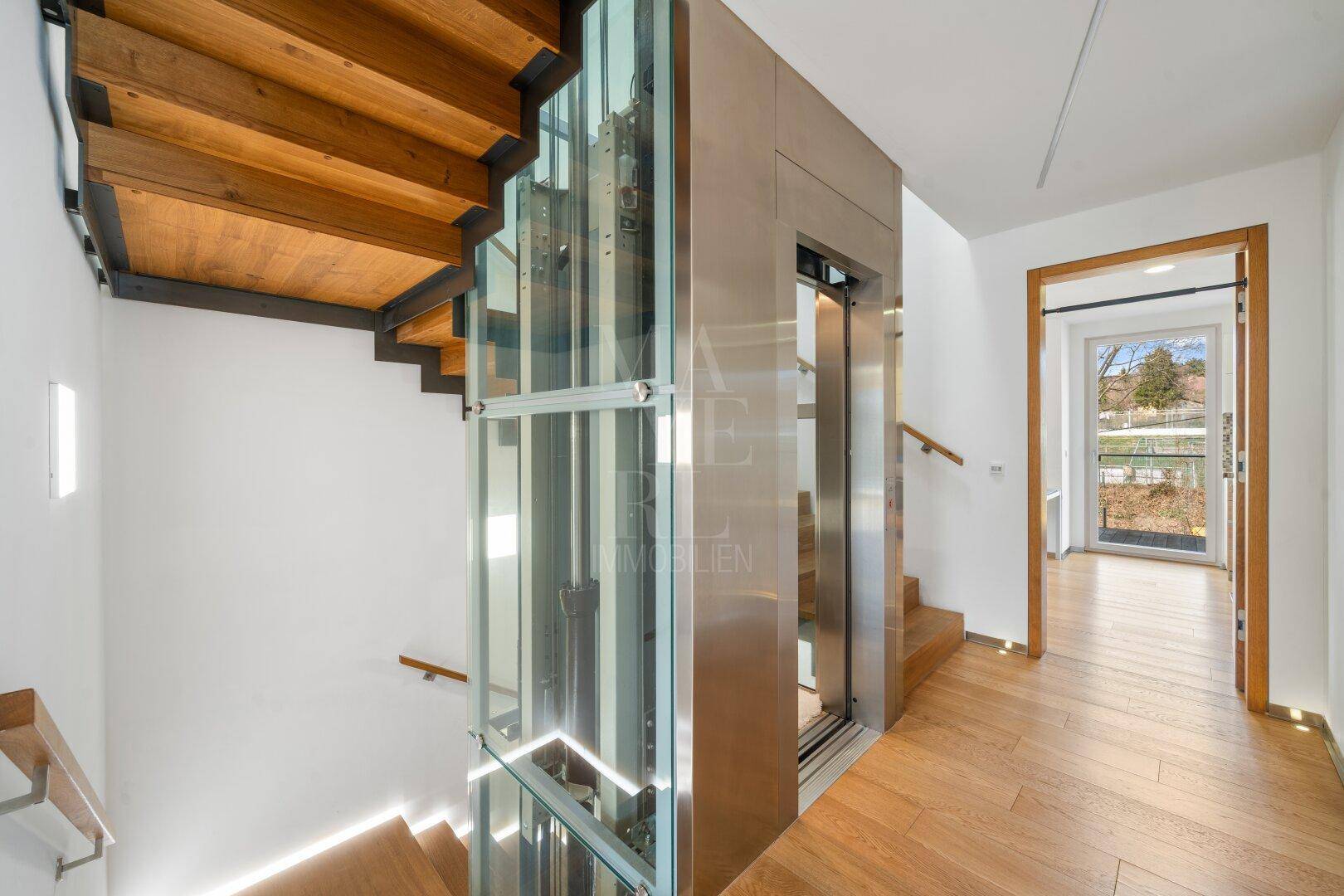 Moderner Eingangsbereich mit Holztreppe und einem eleganten Glaslift, der die Etagen verbindet.