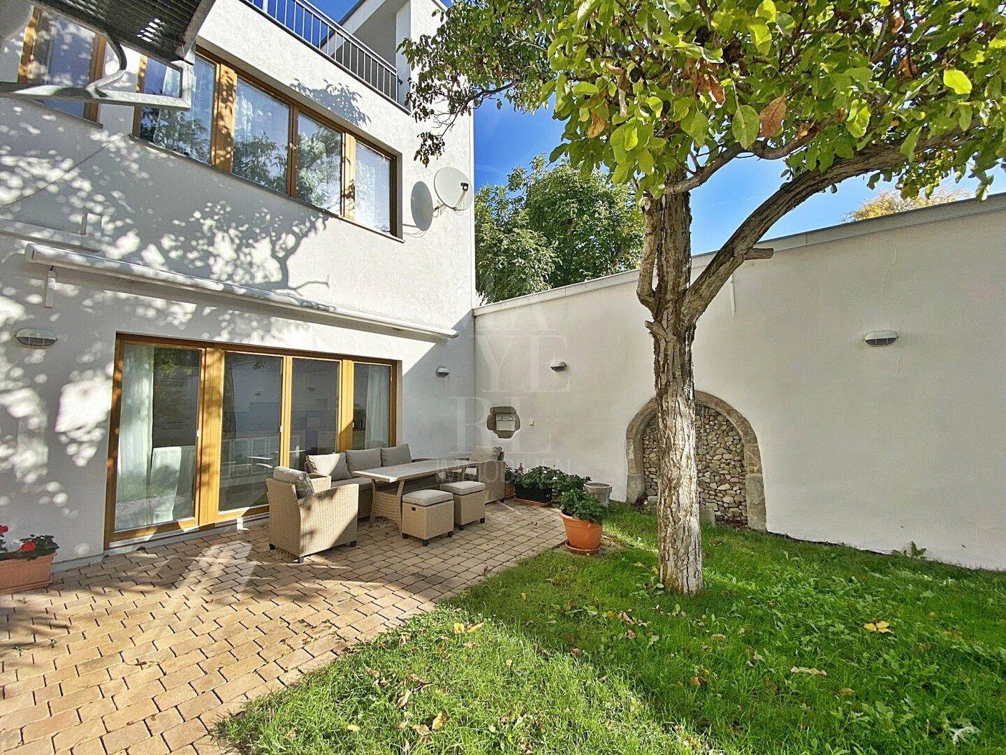 Gemütliche Terrasse mit Sitzgelegenheiten, Rasen und einem alten Steinbogen in der Wand.