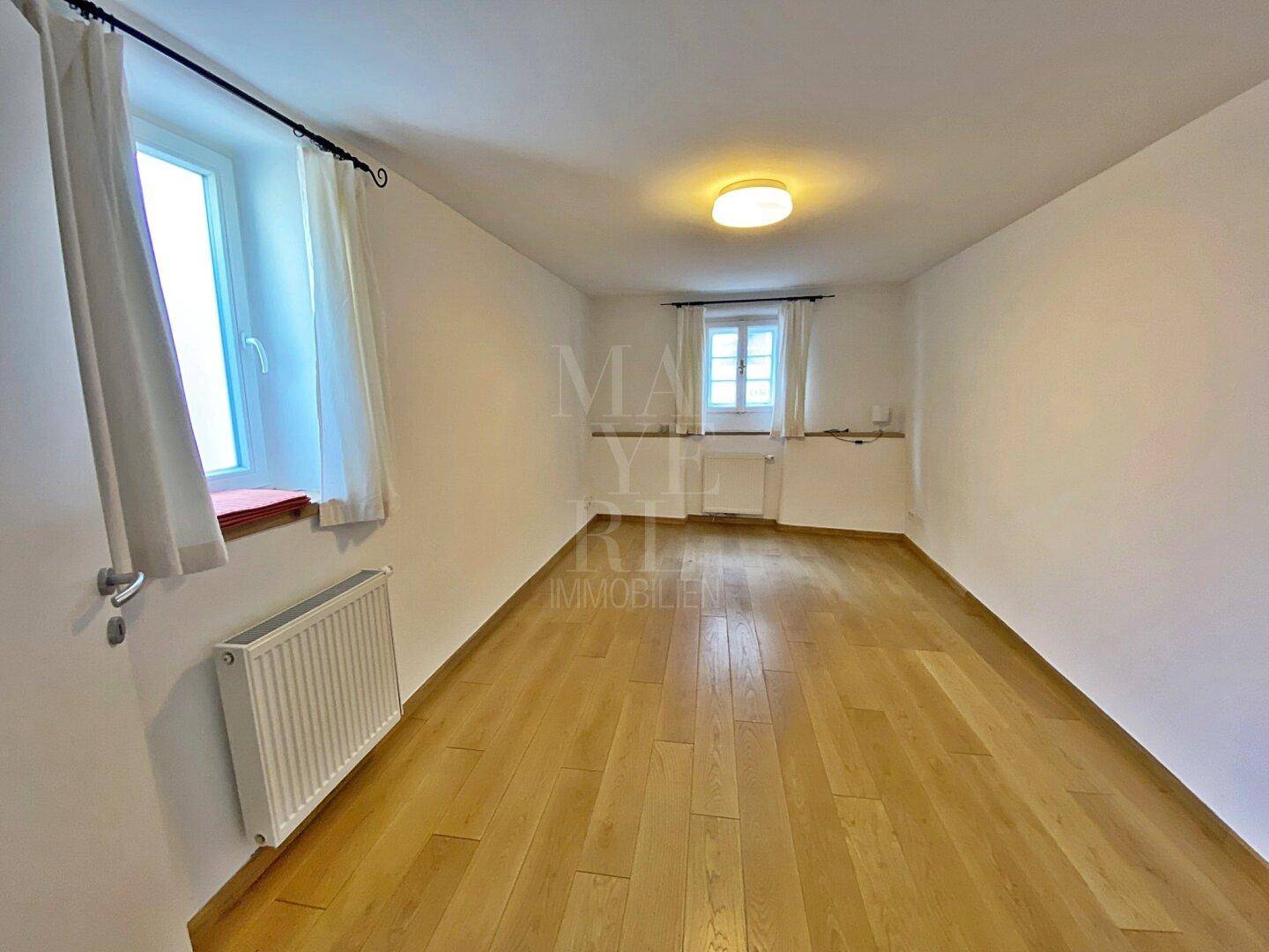 Leeres Zimmer mit Holzboden und zwei Fenstern, bereit zur individuellen Gestaltung.