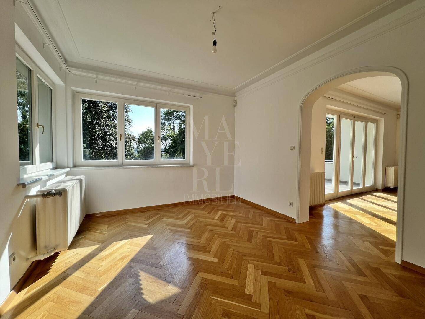Geräumiges Wohnzimmer mit Fischgrätparkett, hohen Decken und Blick ins Grüne durch große Fenster.