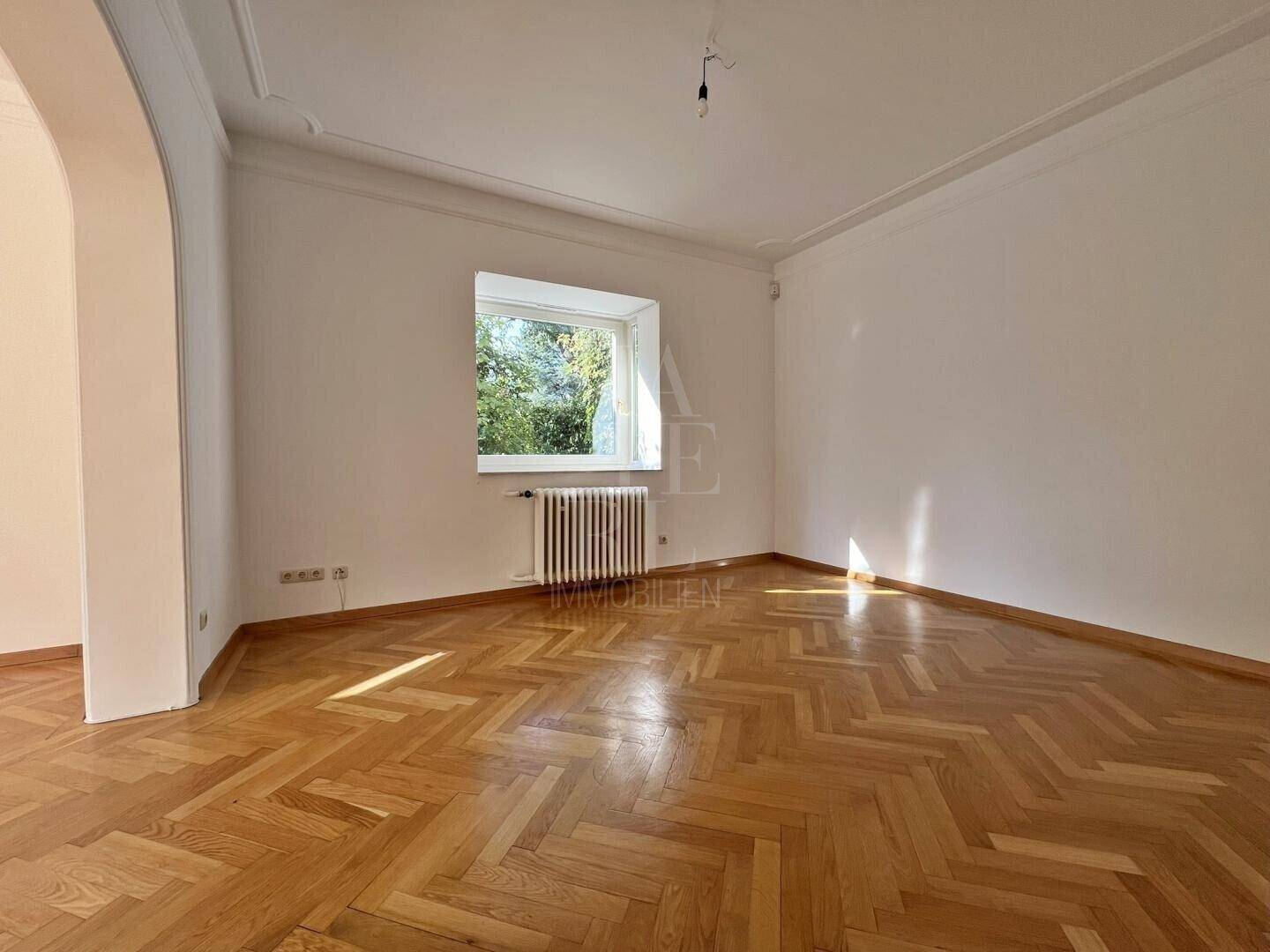 Heller Wohnbereich mit klassischem Fischgrätparkett und einem Fenster mit Blick ins Grüne.