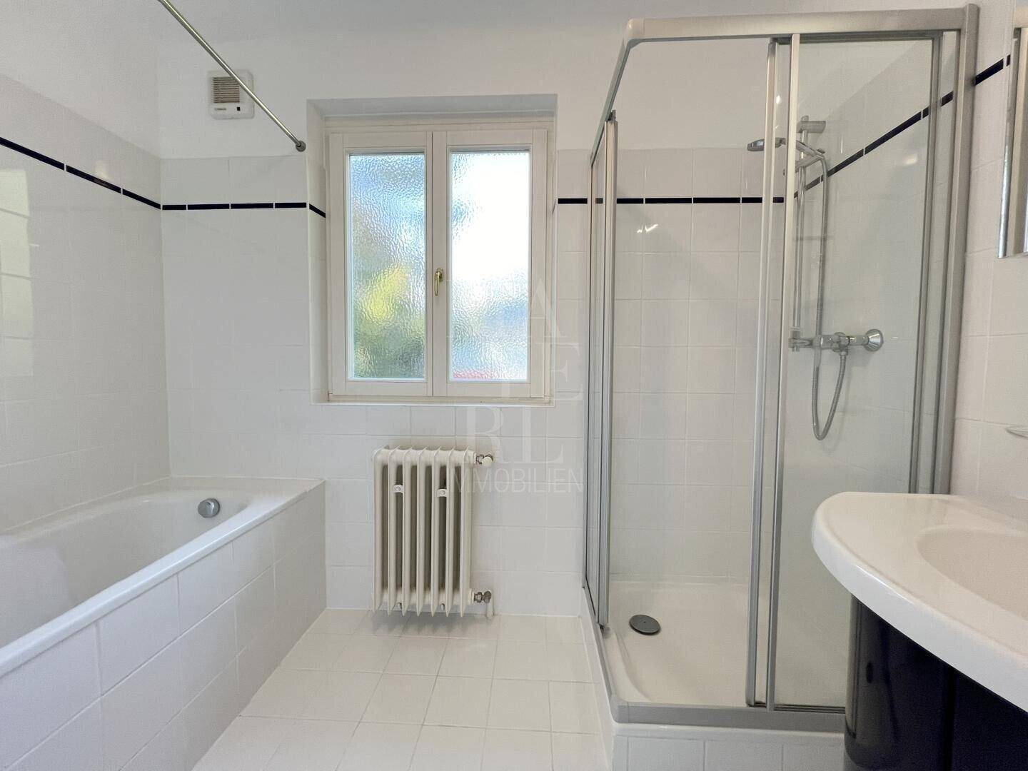 Helles Badezimmer mit weißen Fliesen, Badewanne, Duschkabine und Fenster.