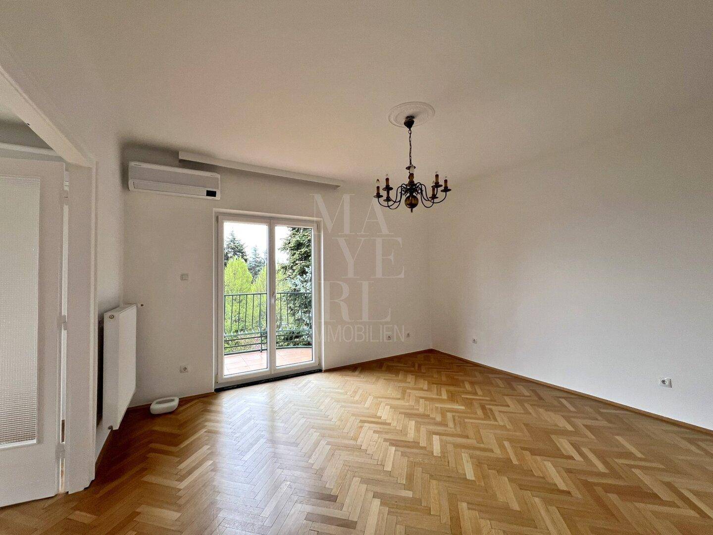 Helles Schlafzimmer mit Fischgrätparkett, Klimaanlage und Zugang zu einem kleinen Balkon mit Grünblick.