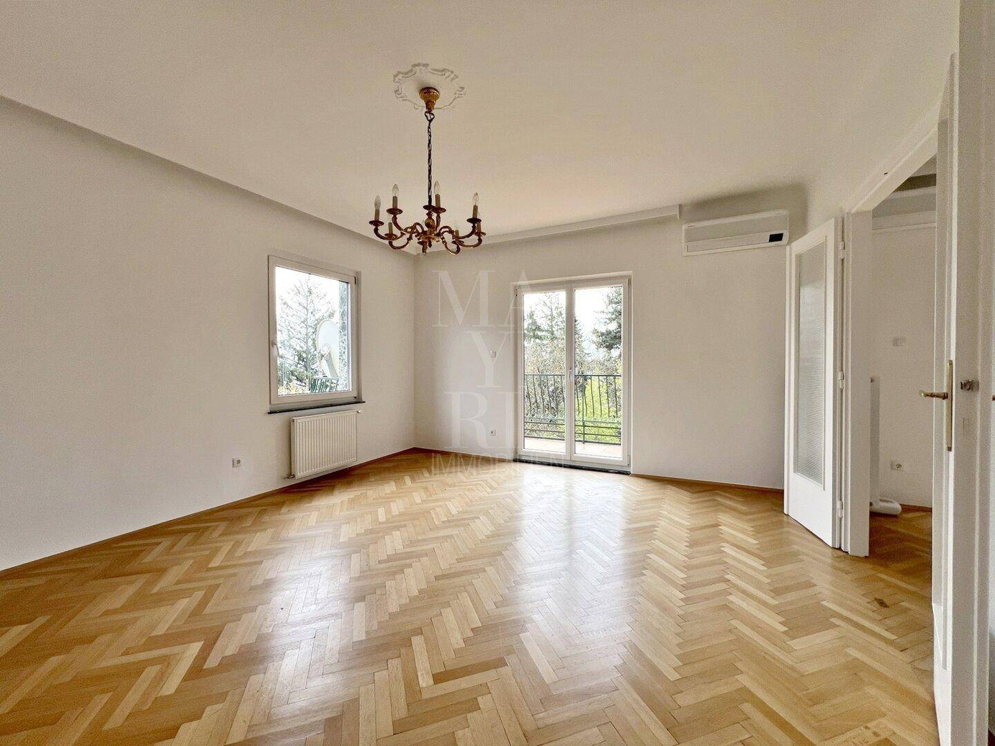 Geräumiges Schlafzimmer mit Fischgrätparkett, Kronleuchter und zwei Fenstern, die viel Licht hereinlassen.