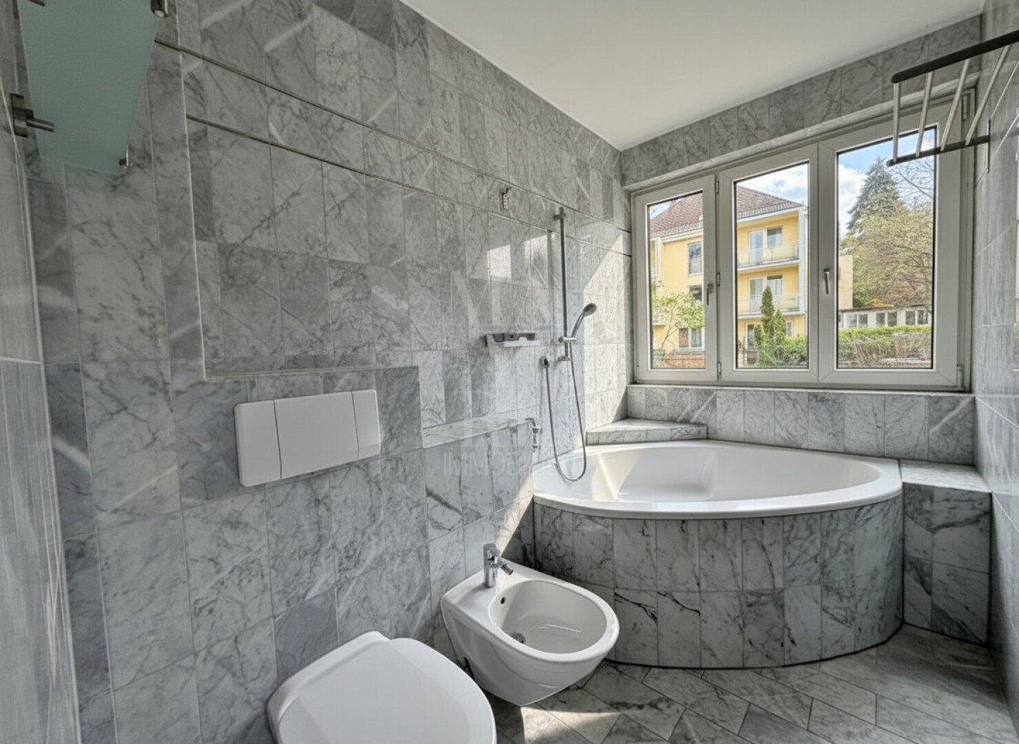 Elegantes Badezimmer mit Marmorfliesen, Eckbadewanne, Toilette und Bidet unter einem Fenster.