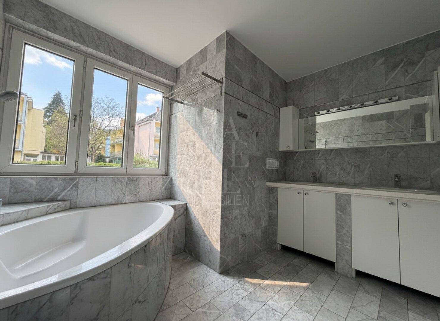 Großes Badezimmer mit grauen Marmorfliesen, Eckbadewanne, Doppelwaschbecken und großem Fenster.