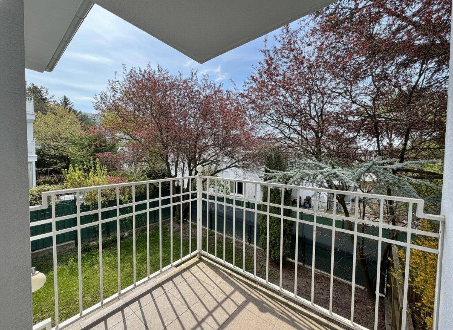 Heller Balkon mit weißem Geländer und Blick auf den grünen Garten und die umliegenden Bäume.