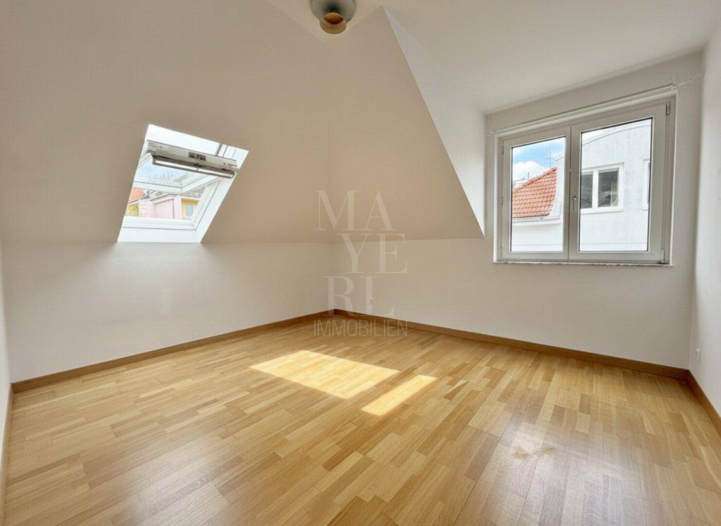 Geräumiges Dachzimmer mit Holzfußboden, viel Tageslicht durch Fenster und Dachschräge.