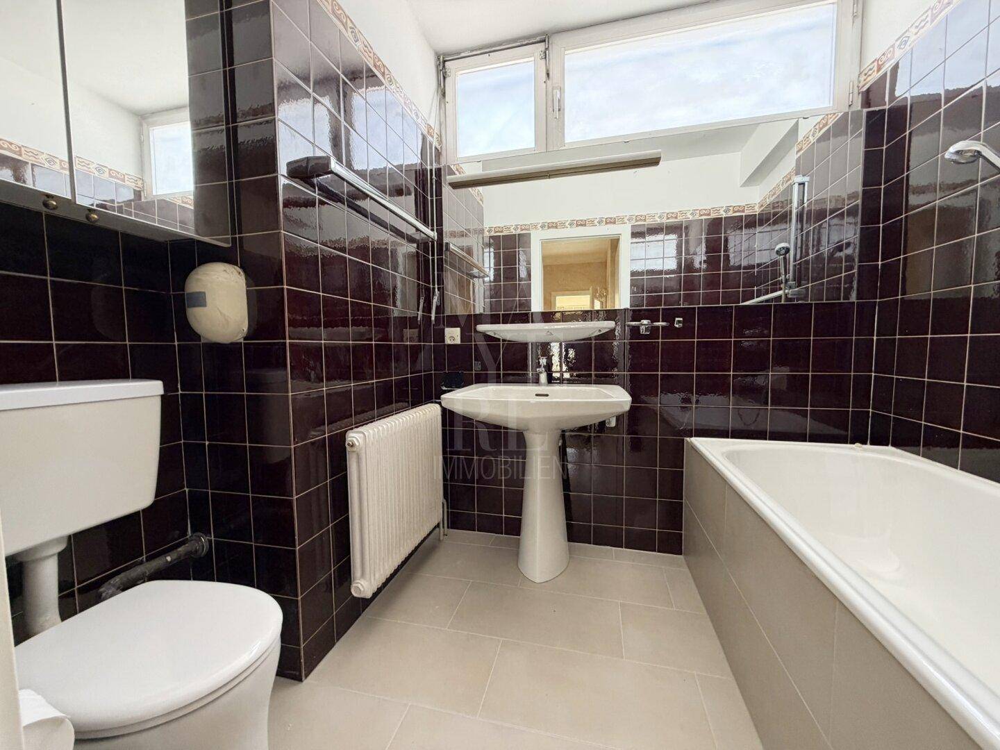 Badezimmer mit dunklen Fliesen, Badewanne, WC und Fenster für Tageslicht.