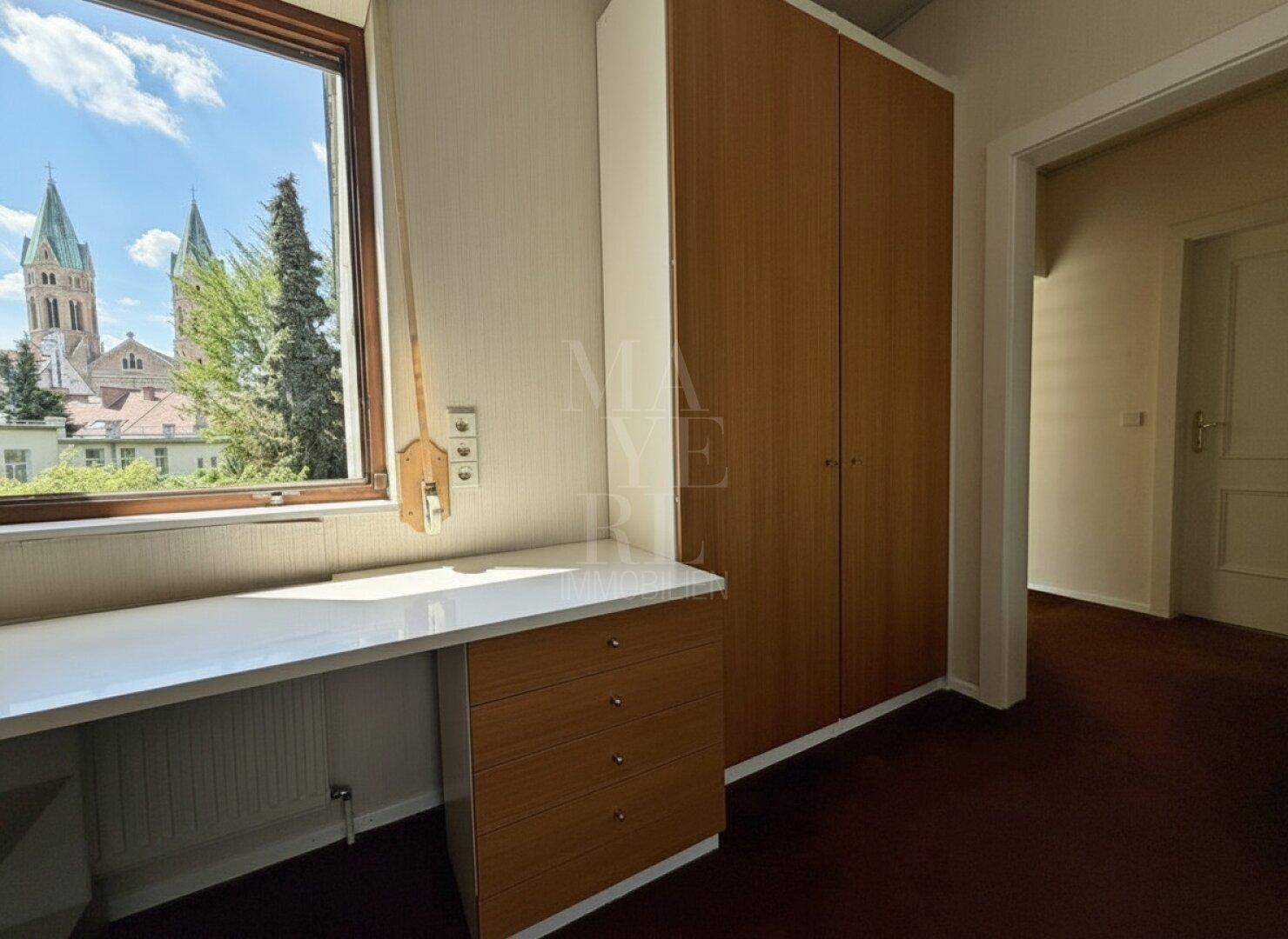 Arbeitszimmer mit Schreibtisch, Einbauschrank und Fenster mit Blick auf die Kirche.