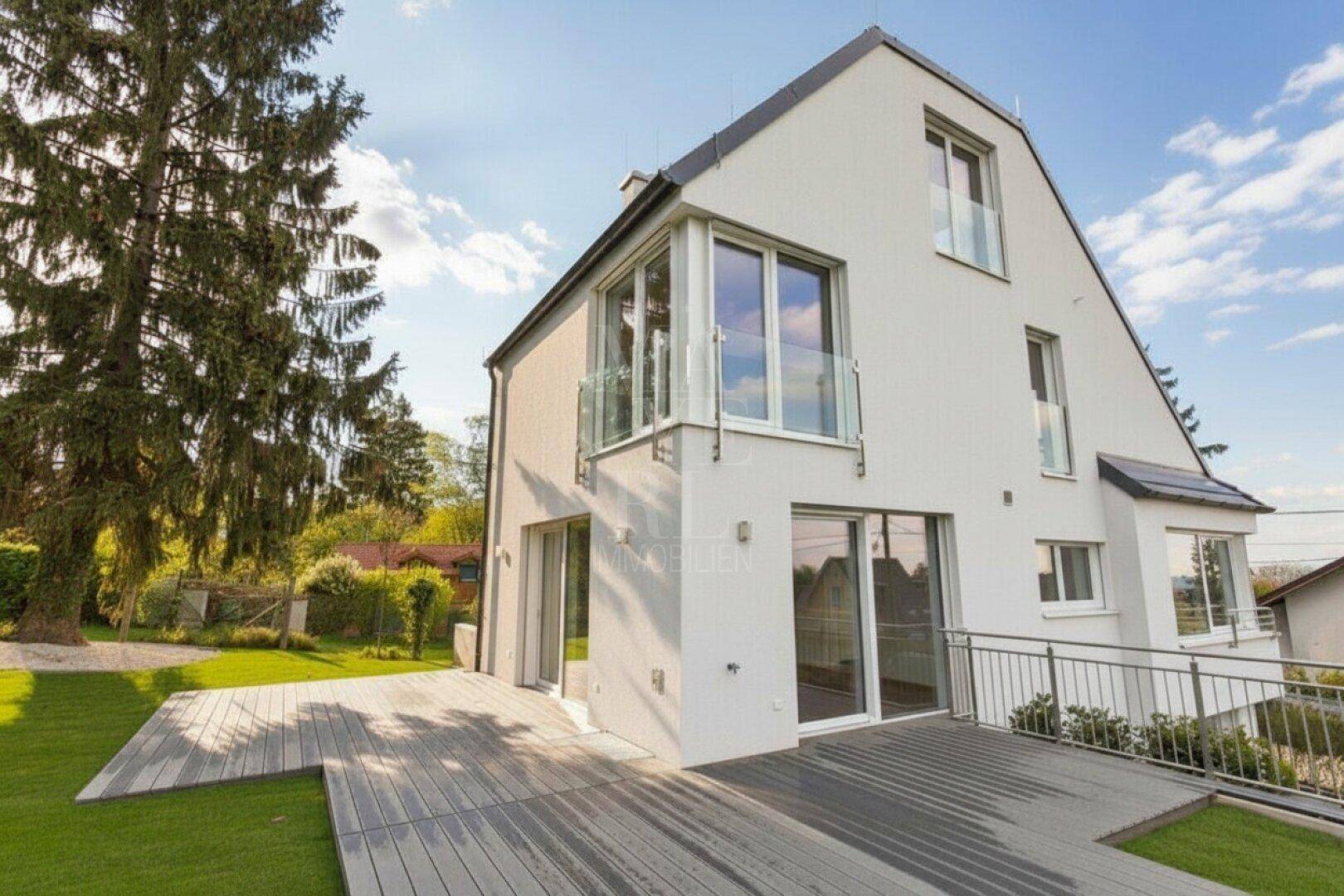 Modernes weißes Einfamilienhaus mit großer Holzterrasse und gepflegtem Garten unter blauem Himmel.