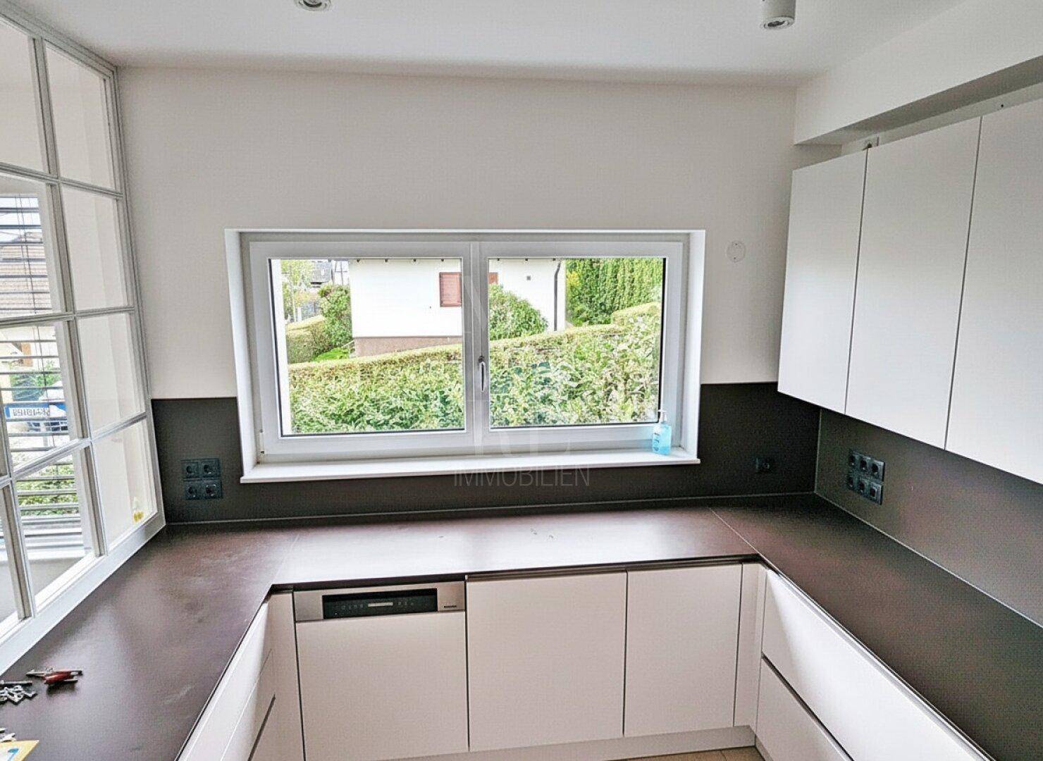 Moderne Einbauküche mit weißen Schränken, dunkler Arbeitsplatte und Fenster mit Blick ins Grüne.