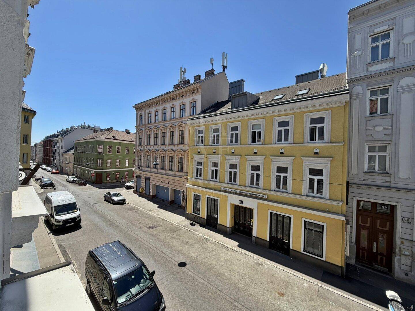 Sonniger Blick auf eine belebte Straße mit historischen Gebäuden und parkenden Fahrzeugen.