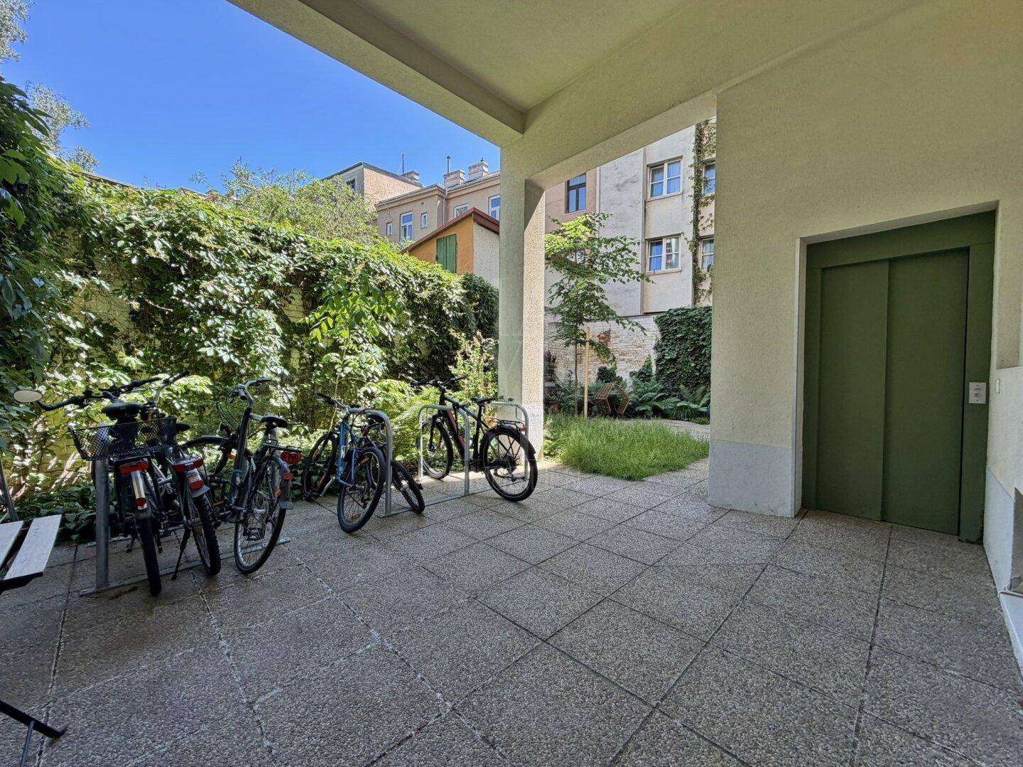 Überdachter Außenbereich mit Fahrradabstellplätzen und Zugang zu einem Aufzug im Gebäude.