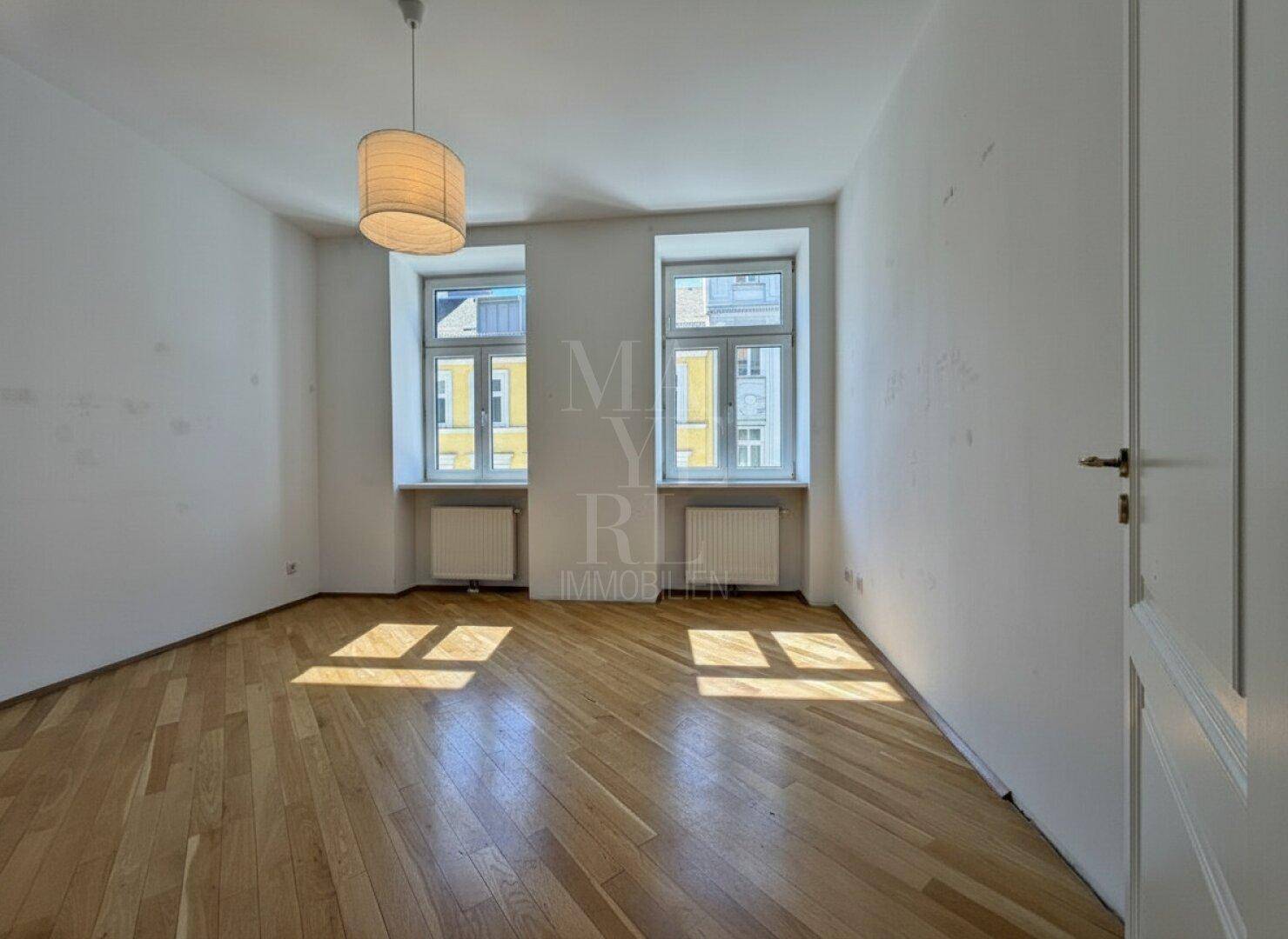 Helles Schlafzimmer mit Parkettboden und zwei Fenstern, die viel Tageslicht hereinlassen.