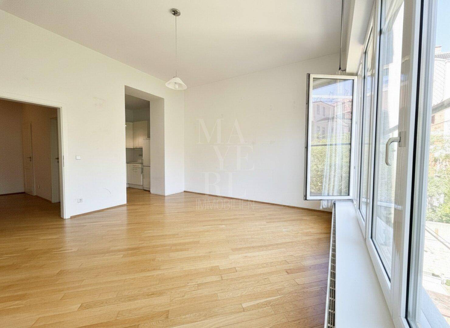 Geräumiges Wohnzimmer mit Parkettboden und großen Fenstern, offen zu einem angrenzenden Bereich.