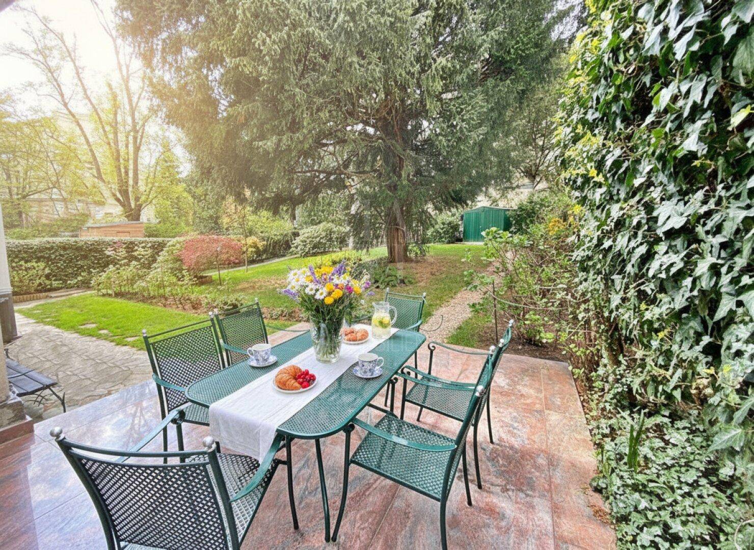 Gemütliche Terrasse mit Esstisch und Stühlen, umgeben von üppigem Grün und einem großen Baum.