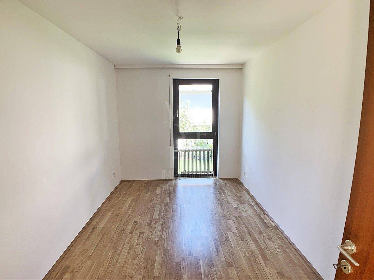 Helles Schlafzimmer mit Parkettboden und großem Fenster, das viel Tageslicht hereinlässt.