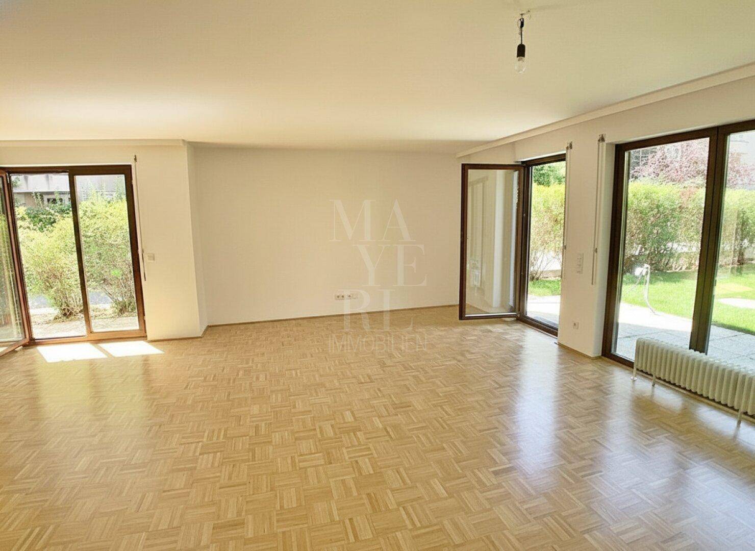 Geräumiges Wohnzimmer mit hellem Parkettboden und großen Fensterfronten, die viel Tageslicht hereinlassen.